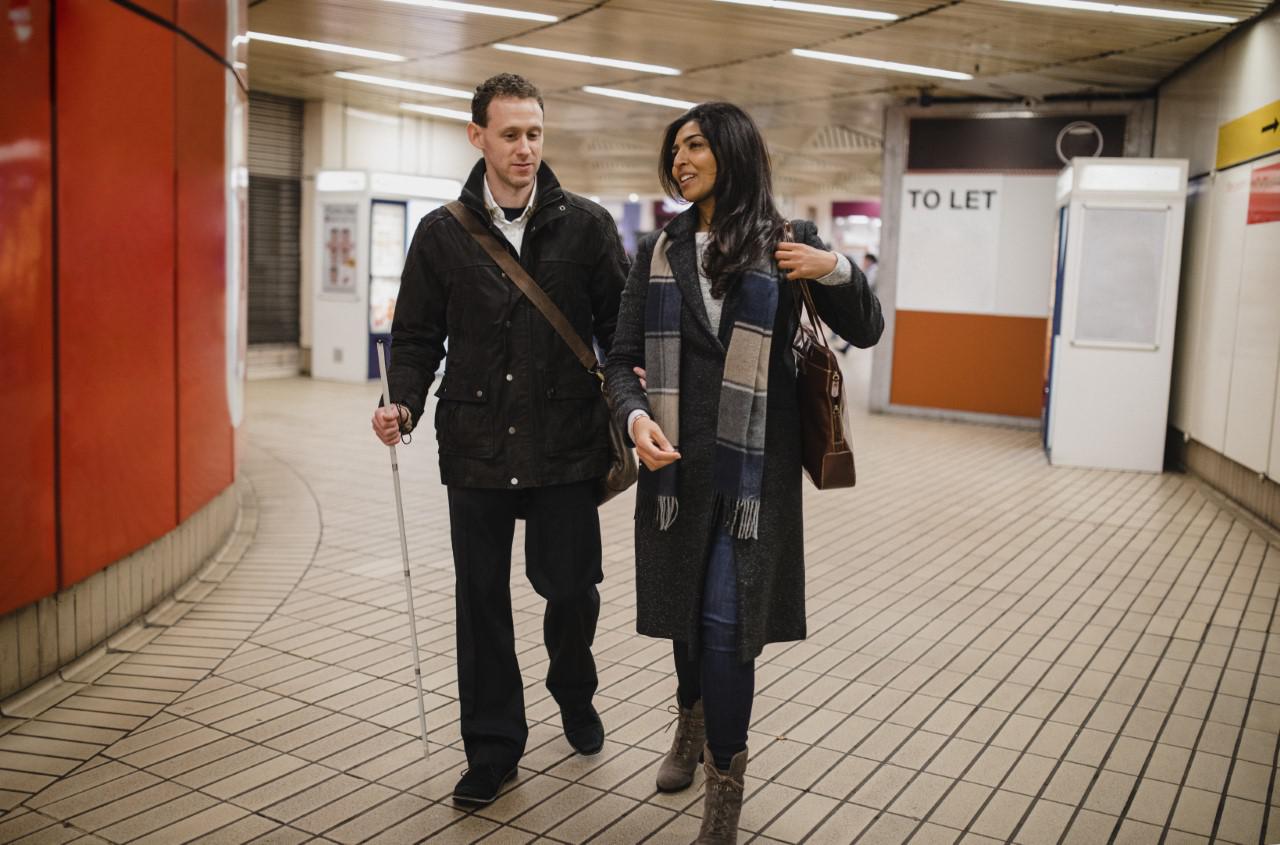 Woman walking with a man in a subway. The man is using a walking cane. 
